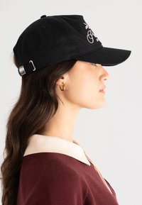 Svart bomullslue med buet skjerm, med hvit brodert tekst. Justerbar sølvspenne på baksiden. Modellen har på seg en burgunder topp.