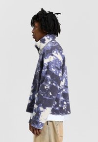 Fleece pullover jacket in purple and white tie-dye pattern. High collar, long sleeves, and boxy fit. Textured, warm fabric with soft surface.