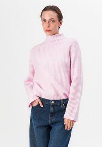 Pulover roz deschis cu guler înalt, cu maneci lungi, asortat cu blugi de denim închis. Puloverul are o croială lejeră și un material texturat.