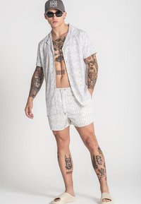 Witte shirt met korte mouwen en bijpassende shorts met een algeheel patroon. Gecombineerd met een zonnebril en een grijze pet, in combinatie met lichte instap-sandalen.