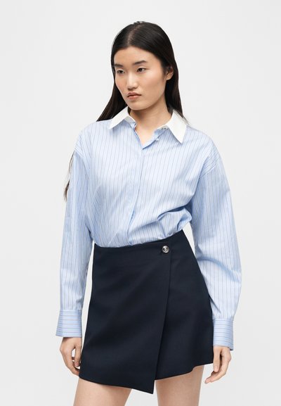 Γυναίκα με μπλε πουκάμισο με ρίγες και λευκό γιακά και μαύρη ασύμμετρη φούστα στέκεται μπροστά σε λευκό φόντο, κοιτώντας ελαφρώς προς τα αριστερά.