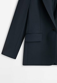 Veste de costume bleu marine foncé à manches longues, fermeture par un bouton unique et poche avant à rabat sur fond uni.