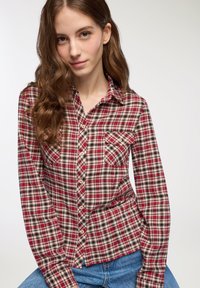 Giovane donna con lunghi capelli castani mossi, che indossa una camicia a quadri rossi e beige e jeans blu, su uno sfondo neutro.