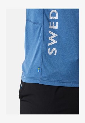 Niebieska koszulka sportowa z napisem "SWED" wydrukowanym pionowo na białym tle. Posiada akcent w postaci flagi szwedzkiej na dole. Gładka tekstura, krótkie rękawy.