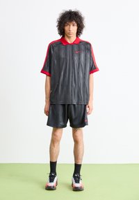 Giovane con capelli ricci che indossa abbigliamento sportivo Nike nero e rosso e sneaker, in piedi su un pavimento verde contro uno sfondo bianco.