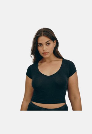 Junge Frau mit langen dunklen Haaren, die ein enganliegendes schwarzes V-Ausschnitt-Crop-Top vor einem schlichten weißen Hintergrund trägt.