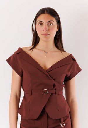 Giovane donna con capelli lisci e scuri che indossa un top marrone scollato con cintura e una collana d'argento con un ciondolo verde.