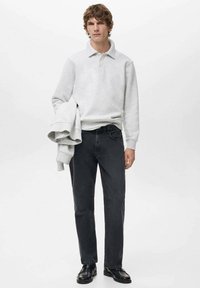 Szary sweter z kołnierzykiem, zestawiony z ciemnymi dżinsami. Model trzyma dopasowaną szarą bluzę. Czarnie buty dopełniają całość.