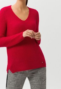 Pull en tricot rouge avec un décolleté en V, des manches longues et des fentes sur les côtés, associé à un pantalon à carreaux avec un motif texturé.