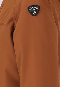 Bruine buitenjas met een gestructureerd oppervlak, voorzien van een zwart logo met de "Zigzag" op de linkerschouder.