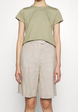 Person wearing olive green short-sleeve t-shirt tucked into high-waisted, beige, knee-length tailored shorts against a plain background.