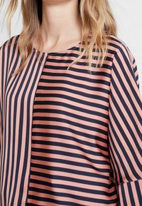 Blouse rayée aux couleurs bleu marine et rose pâle, en tissu lisse, avec un col arrondi et une coupe ample à manches trois-quarts.