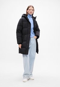 Cappotto puffo nero con cappuccio, caratterizzato da un design trapuntato. Indossato sopra un dolcevita azzurro chiaro e abbinato a un paio di jeans a gamba larga azzurro chiaro e scarpe bianche.