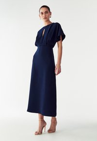 Laivansinis midi-mekko, jossa on lyhyet, volyymikkaat hihat ja avointa muotoa kaula-aukolla. Silkkinen kangas, suora siluetti, yhdistettynä nude-korkokenkiin.