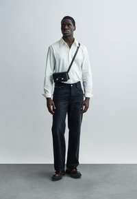 Λευκό πουκάμισο με κουμπιά, σκούρο τζιν, μαύρη τσάντα ώμου με μεταλλικά στοιχεία και σκούρα παπούτσια. Στέκεται μπροστά σε ουδέτερο φόντο.