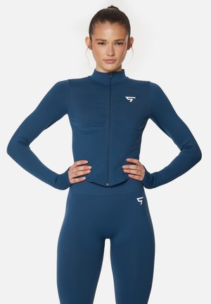 Chaqueta deportiva azul marino con cierre y mangas largas, con un diseño cropped, combinada con leggings de talle alto a juego. Cuenta con acentos de logo en blanco.