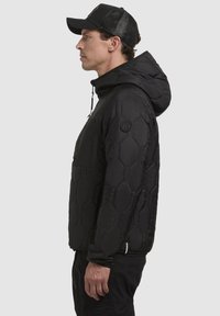 Schwarze Kapuzenjacke mit einem sechseckigen Steppmuster, elastischem Saum, Kordelzugkapuze und einer Taschenfront. Aus synthetischem Material gefertigt.