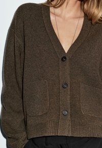 V-hals, bruine geribde cardigan met lange mouwen, voorzien van zwarte knopen en twee voorzakken. Zachte textuur en een comfortabele pasvorm.
