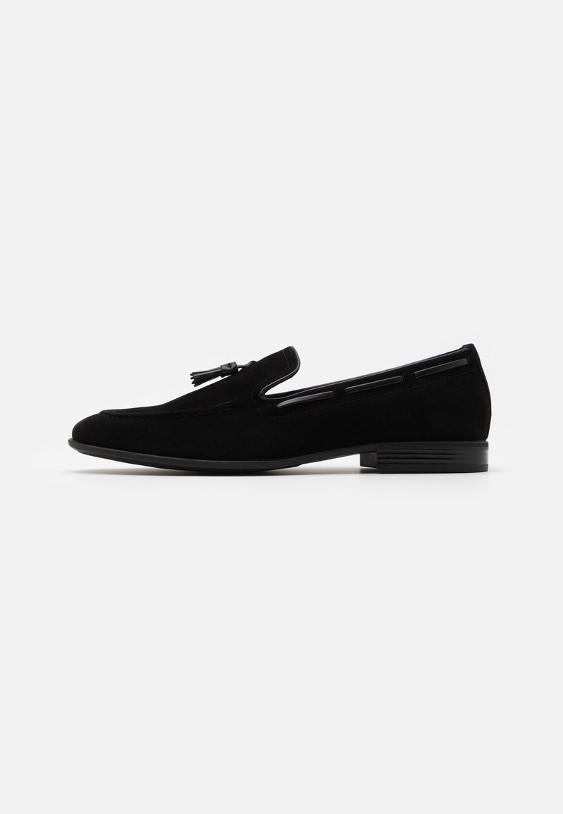 Mocassin homme en daim noir avec détail pompon, petit talon empilé et coutures discrètes sur fond blanc.