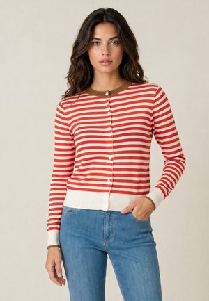 Donna con lunghi capelli scuri che indossa un cardigan a righe orizzontali rosse e bianche e jeans blu, in piedi davanti a uno sfondo uniforme.