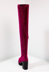 Botte en velours bordeaux haute avec un talon bloc, vue de l'arrière, posée sur une surface blanche contre un fond blanc uni.