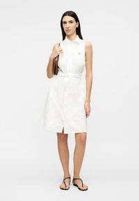 Nő, fehér, ujjatlan, derékban öves, galléros ruhát visel, barna táskát tart a vállán, semleges arckifejezéssel áll, fekete papuccsal.