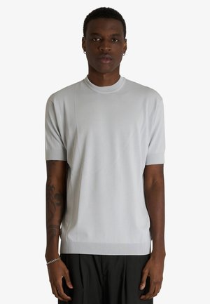 Giovane uomo che indossa una maglietta di maglia a maniche corte grigio chiaro e pantaloni neri, in piedi con le braccia rilassate davanti a uno sfondo bianco uniforme.