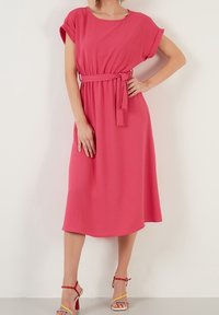 Rosa midi-klänning med korta ärmar och midjesnöre. Mjuk, strukturerad tyg; lätt utsvängd fåll. Kombinerad med flerfärgade klacksandaler.
