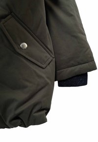 Donkergroen gewatteerde jacksleeve met een klepzak, beveiligd met een zilveren drukknoop en een zichtbare marineblauwe ribbelmanchet.
