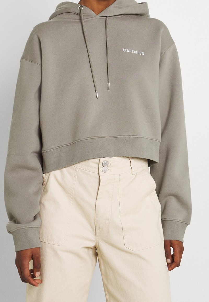 Sudadera gris corta de tejido suave con capucha ajustable y logo blanco. Combinada con pantalones de color claro que tienen dos bolsillos frontales.