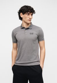 Pilkas polo marškinėliai iš minkštos medžiagos, su apykakle, trumpomis rankovėmis ir mažu EA7 logotipu ant krūtinės. Dėvimi su juodomis kelnėmis.