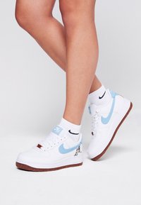 Benen in witte Nike Air Force 1 sneakers met lichtblauwe swoosh, bruine zolen en witte Nike sokken met zwarte swoosh op een neutrale achtergrond.