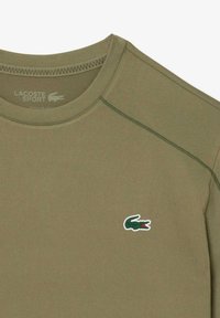Cămașă scurtă de culoare verde măsliniu, dintr-un material neted, cu gât rotund. Prezintă un logo brodat în formă de crocodil, de culoare verde, pe partea stângă a pieptului.