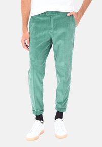 Pantaloni in corduroy verde con una vestibilità affusolata, dotati di chiusura a bottone, tasche laterali e orli arrotolati. Indossati con sneakers bianche.