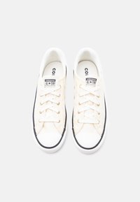 Ζευγάρι λευκά παπούτσια Converse All Star χαμηλά, με λευκά κορδόνια, μαύρη επένδυση και υφή υφασμάτων στο πάνω μέρος, από επάνω σε λευκό φόντο.