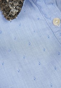 Camicia azzurro chiaro con un tessuto texturizzato che presenta piccoli motivi in rilievo e un colletto decorativo di paillettes. Chiusura a bottoni al collo.