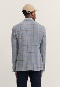 Kariertes Sakko in Blau- und Brauntönen, aus strukturiertem Stoff, mit Revers, Schlitz hinten und knöpfbaren Manschetten. Casual-Cap getragen.
