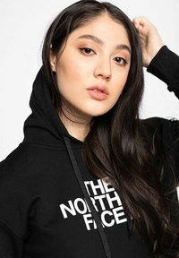 Sudadera negra con un bolsillo canguro en el frente, que presenta un gran logotipo blanco con el texto "THE NORTH FACE" en el pecho y cordones en la capucha.