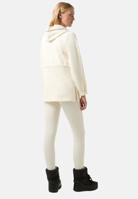 Kremowa kurtka z kapturem z bocznymi rozcięciami i długimi rękawami, w połączeniu z kremowymi legginsami i czarnymi sznurowanymi butami. Gładka tekstura na całej powierzchni.