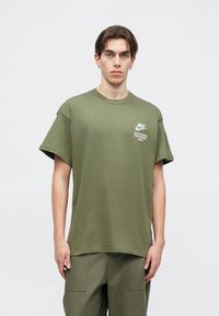 Ελιξίδενη πράσινη βαμβακερή μπλούζα με κοντό μανίκι, στρογγυλή λαιμόκοψη και εκτυπωμένο λογότυπο στο στήθος. Χαλαρή εφαρμογή, συνδυασμένη με αντίστοιχες cargo παντελόνες.