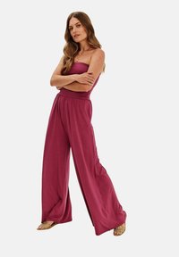 Bordó overal s širokými nohavicemi vyrobený z měkké tkaniny, s bezramínkovým designem, elastickým pasem a postranními rozparky, doplněný o zlaté sandály.