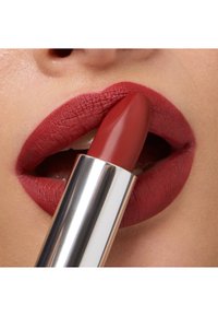 Czerwony matowy pomadka do ust o gładkim wykończeniu trzymana pomiędzy wargami. Tubka ma błyszczące srebrne etui i spiczasty kształt.