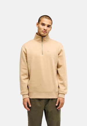 Junger Mann mit kurzen Haaren, der einen beigen Viertel-Zip-Pullover und olivgrüne Hose trägt, steht vor einem schlichten hellen Hintergrund.