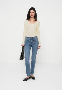 Ανοιχτό κίτρινο μακρυμάνικο τοπ, μπλε τζιν στενής γραμμής, μαύρα μπαλαρίνες, κρατώντας μια μαύρη τσάντα με πουά. Ουδέτερο φόντο.