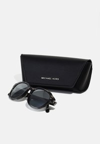 Ochelari de soare Michael Kors cu lentile gri și un cadru translucid, lângă un toc din piele neagră cu un logo argintiu.