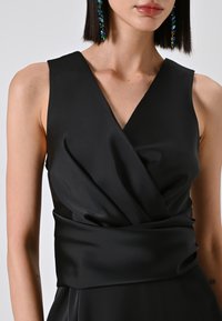 Schwarzes ärmelloses Kleid aus glattem Stoff, mit V-Ausschnitt und gewickeltem Taillen-Detail, ergänzt durch lange, farbenfrohe Ohrringe.
