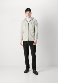 Felpa grigia chiara con zip e tasca frontale, abbinata a pantaloni neri e scarpe nere. L'outfit ha un aspetto casual e stratificato.