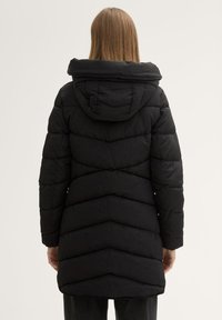 Manteau en duvet noir avec capuche amovible, présentant un motif matelassé en chevrons, des manches longues et un design mi-long.