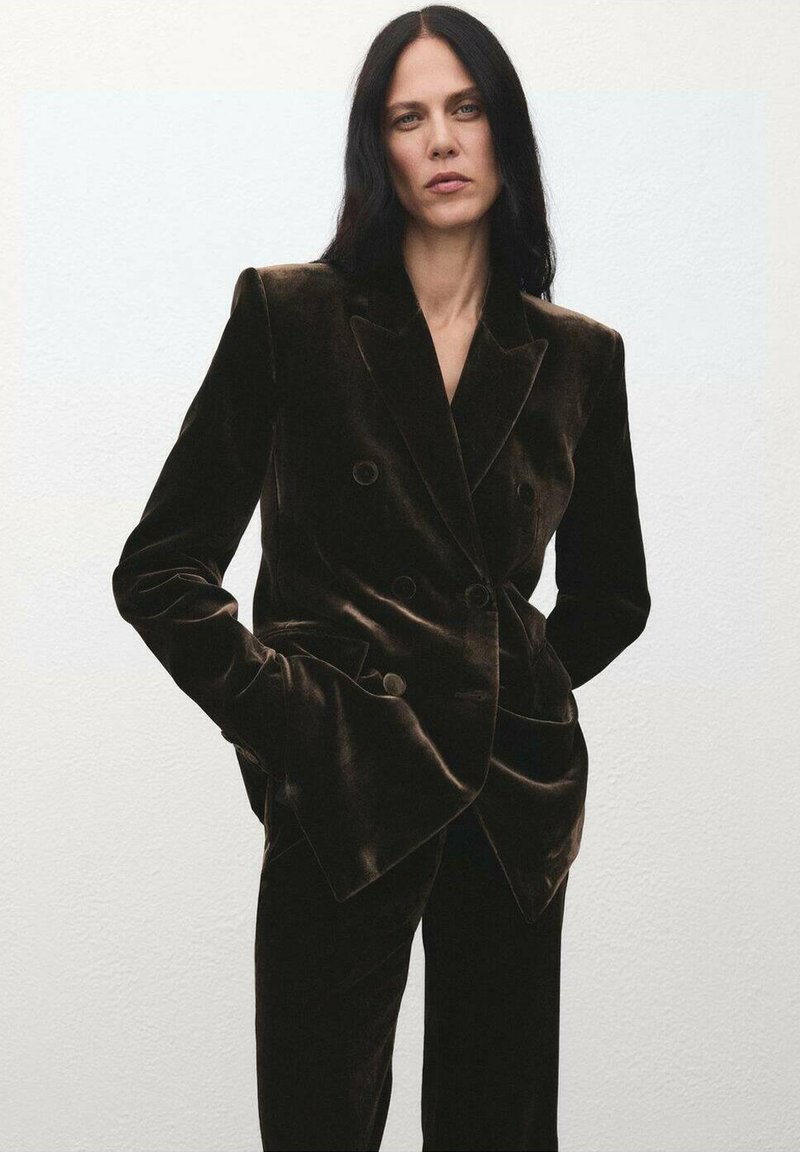 Dvojradový tmavohnedý zamatový sako so širokými klopami, veľkými gombíkmi a štruktúrovaným strihom. Hladká textúra, kombinované s ladiacimi nohavicami.