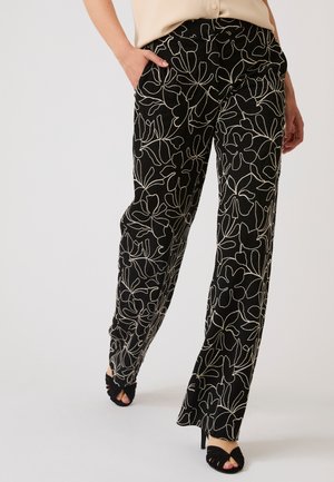 Pantaloni neri a gamba larga con motivo floreale bianco a contorno, indossati con top beige e sandali neri open toe con tacco.